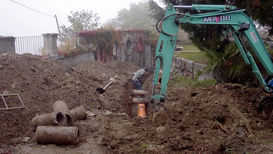 Escavatore al lavoro in uno scavo con tubazioni nel terreno accanto alle murature del giardino.